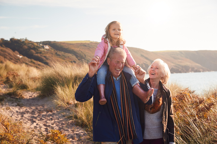 Grandfather Giving Granddaughter Ride On Shoulders As They Walk Through Sand Dunes With Grandmother